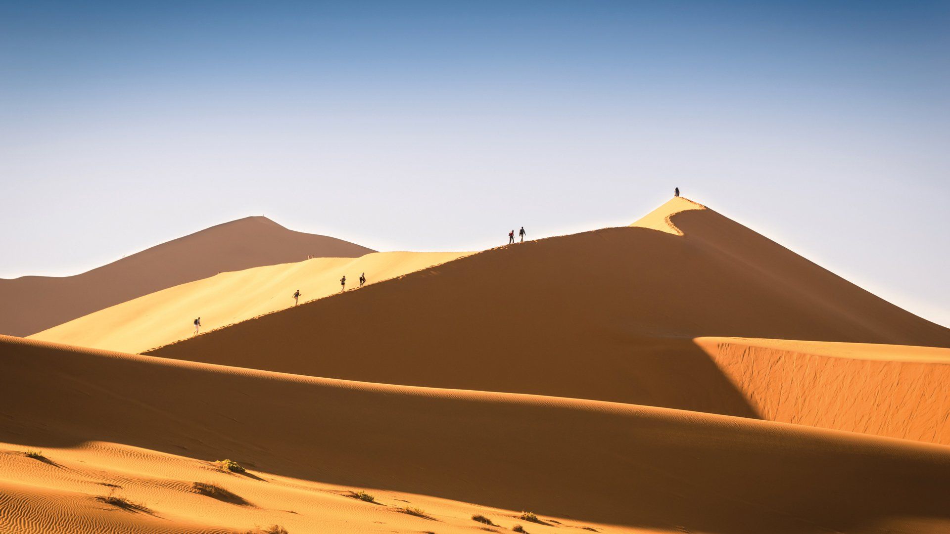 Small figures of people climb a steep, golden sand dune under a clear blue sky.