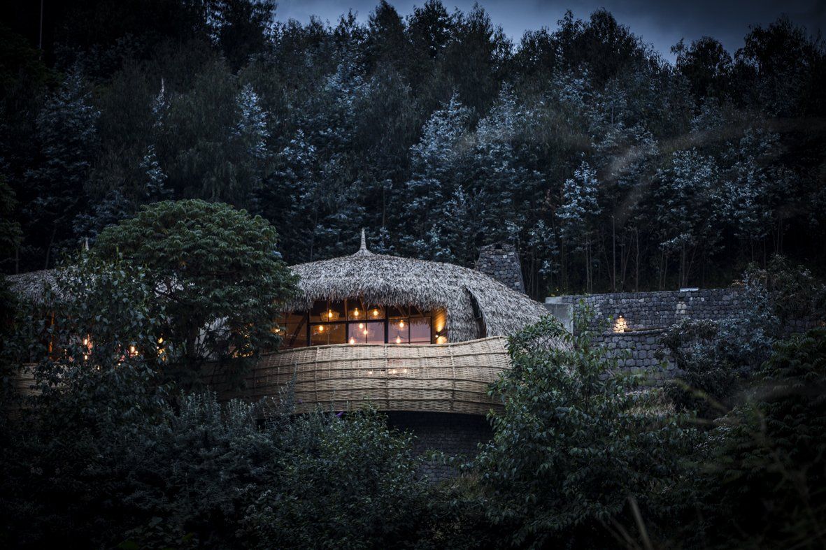 A modern, earth-toned house with a thatched roof glows warmly amid a dark, misty forest at twilight.