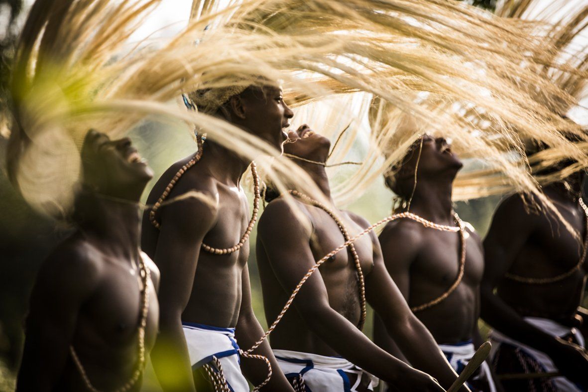 Dancers in traditional attire performing with flowing, straw-like headpieces that arc upwards against a soft-focus backdrop.