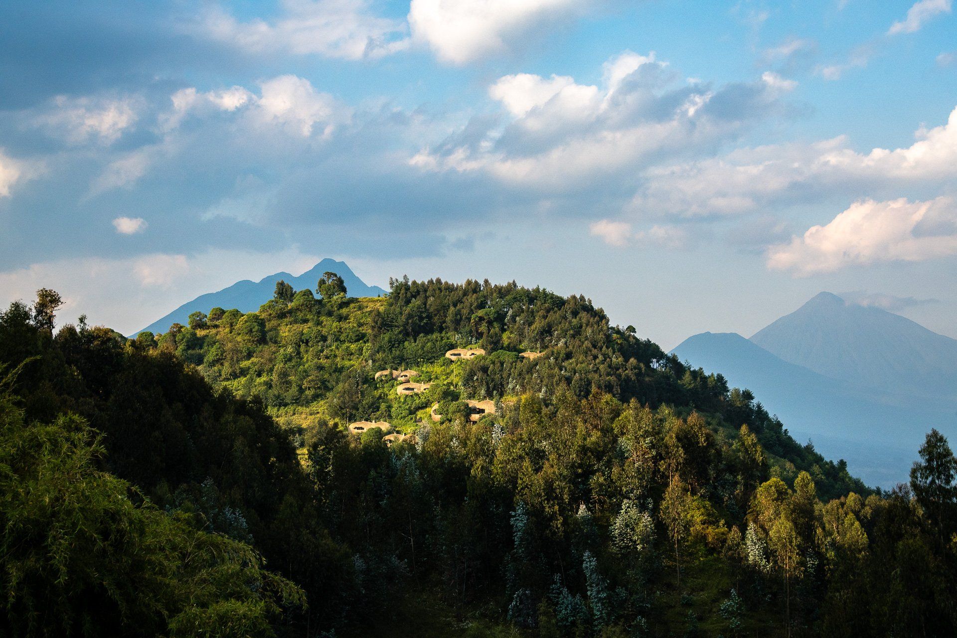 A forested hill under a blue sky with soft, scattered clouds, flanked by distant mountain peaks in the background.