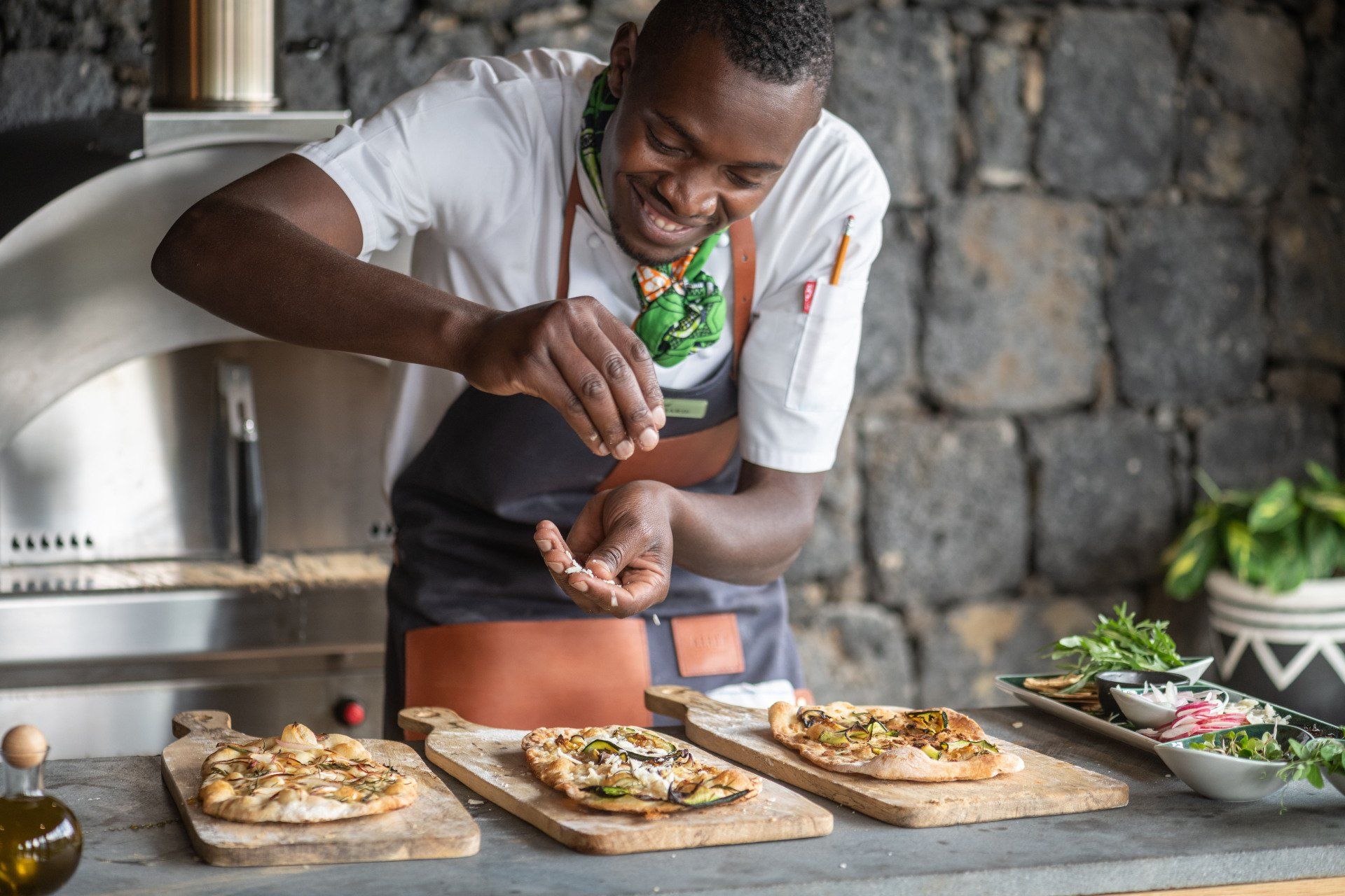 A chef in a white uniform and apron seasons freshly baked flatbreads on wooden boards in a kitchen with a stone wall.