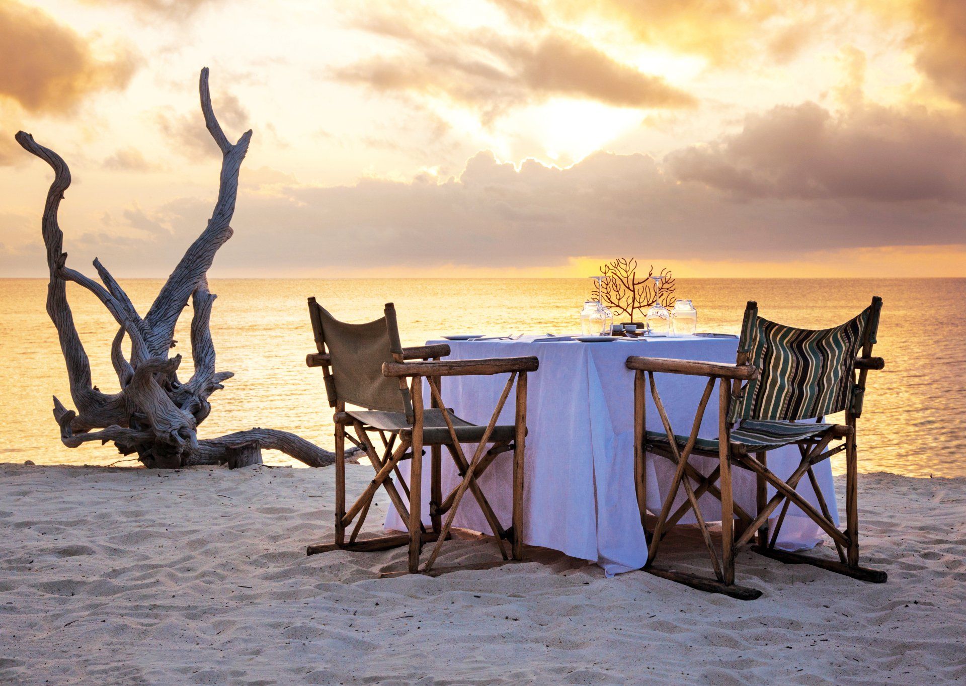 A table set for two on a sandy beach at sunset, with two wooden safari-style chairs and a large piece of driftwood nearby.