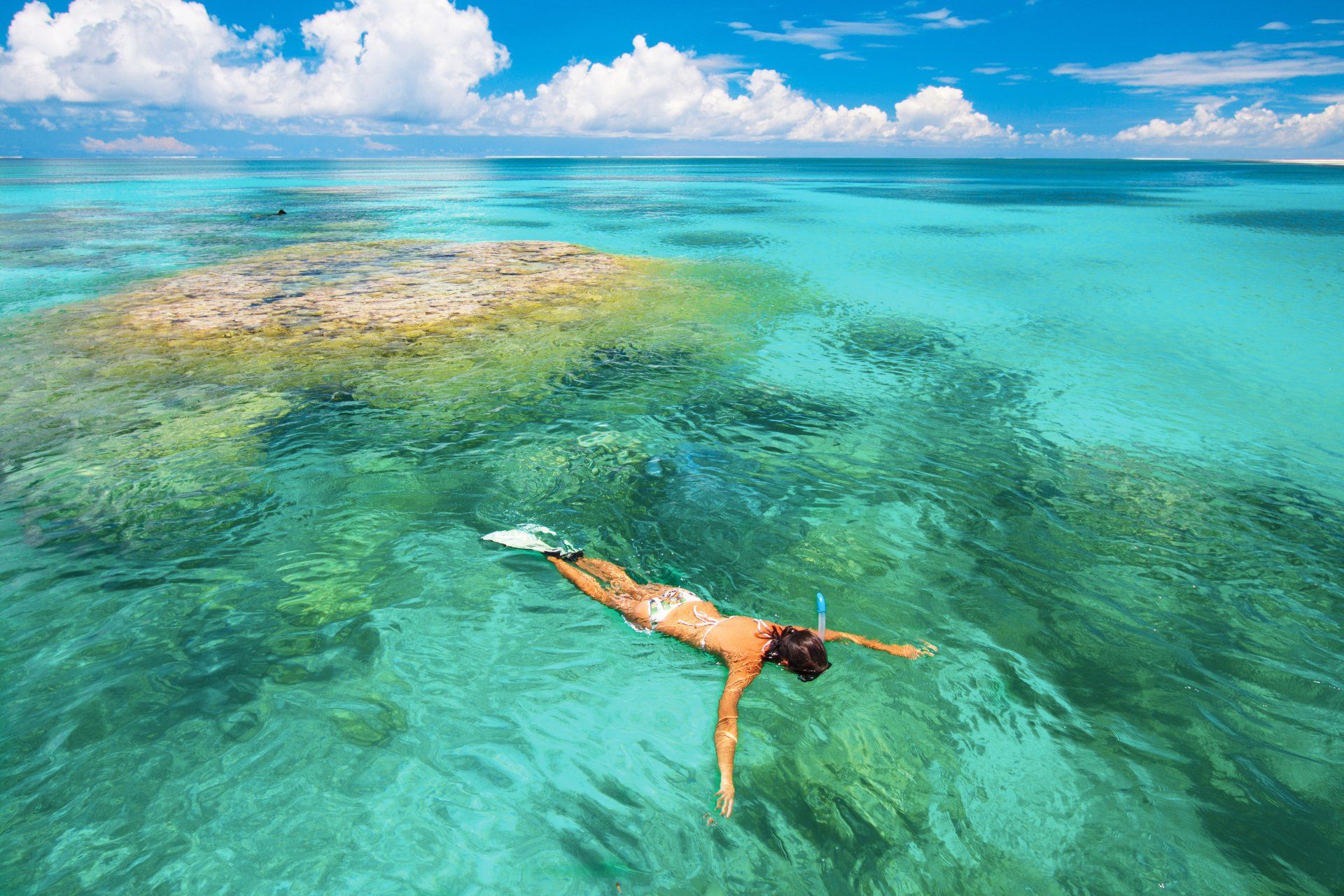 A person snorkels in shallow, crystal-clear turquoise water above a coral reef under a bright, cloudy blue sky.