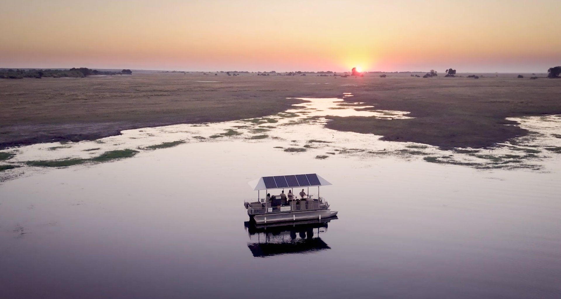 A small, canopy-covered boat carrying people travels across a calm, reflective river at sunset.