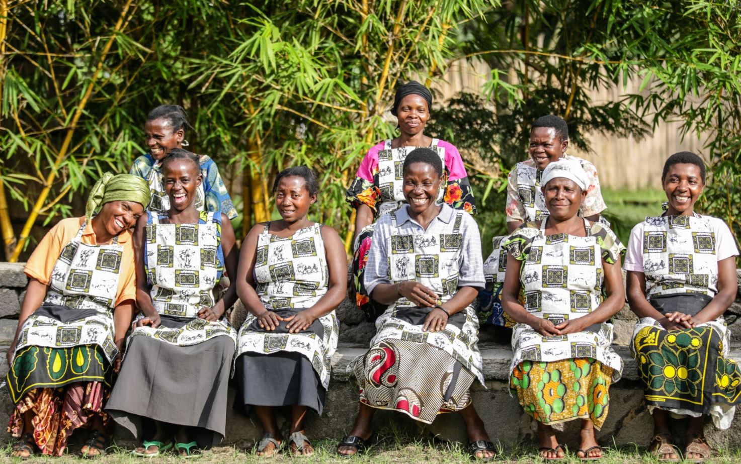 A group of people wearing matching patterned aprons sit in two rows outdoors in front of a bamboo grove.