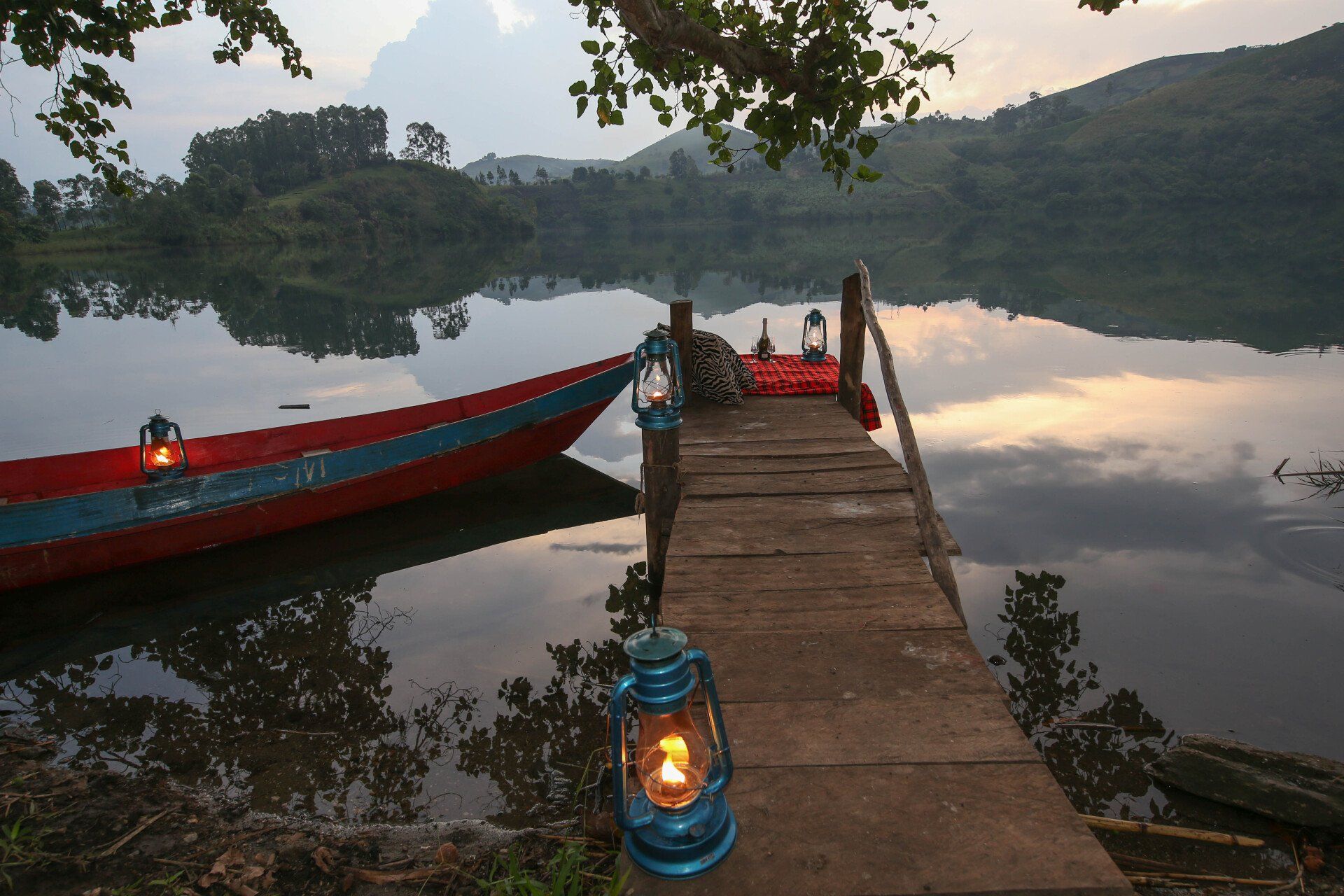 A wooden dock extends into a calm, reflective lake at dusk, illuminated by blue lanterns, with a boat moored alongside.