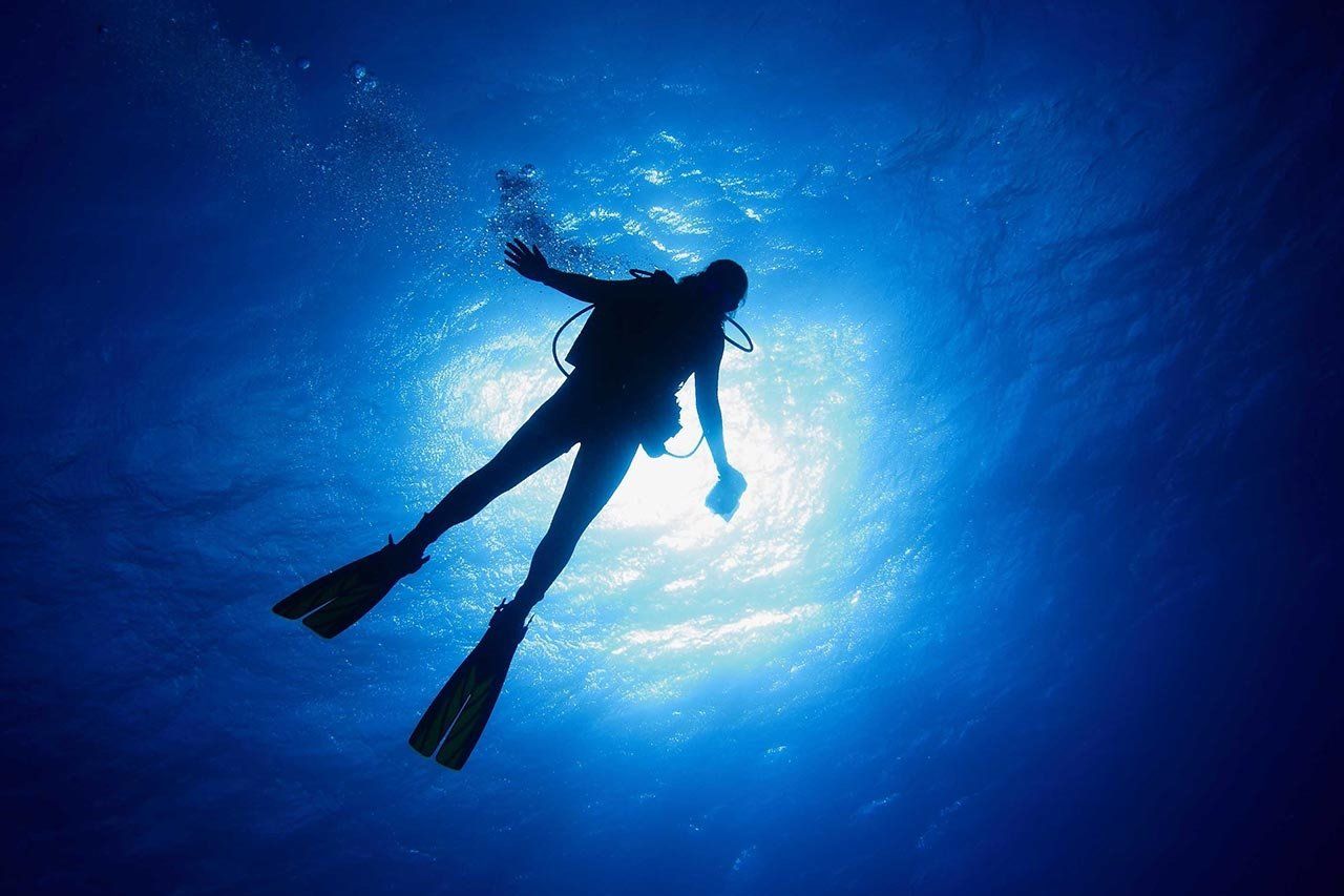 A silhouette of a scuba diver floating underwater, backlit by bright sunlight filtering through the deep blue ocean surface.