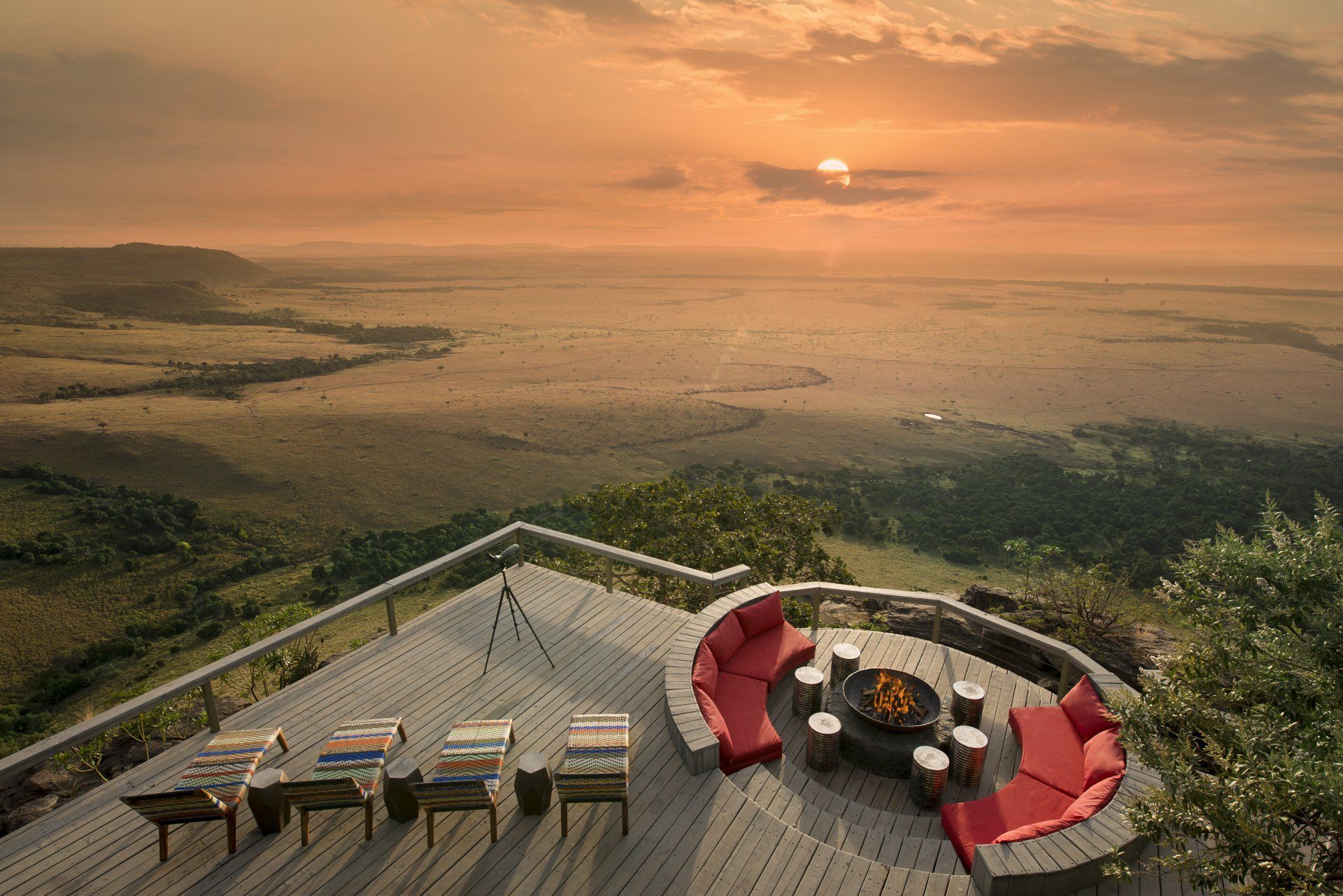 A wooden deck overlooks a vast savanna at sunset, featuring a circular red sofa around a fire pit and several lounge chairs.
