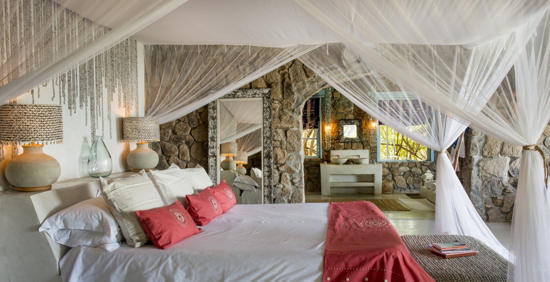A bedroom with stone walls, a bed featuring white and red bedding under a mosquito net canopy, and two bedside lamps.
