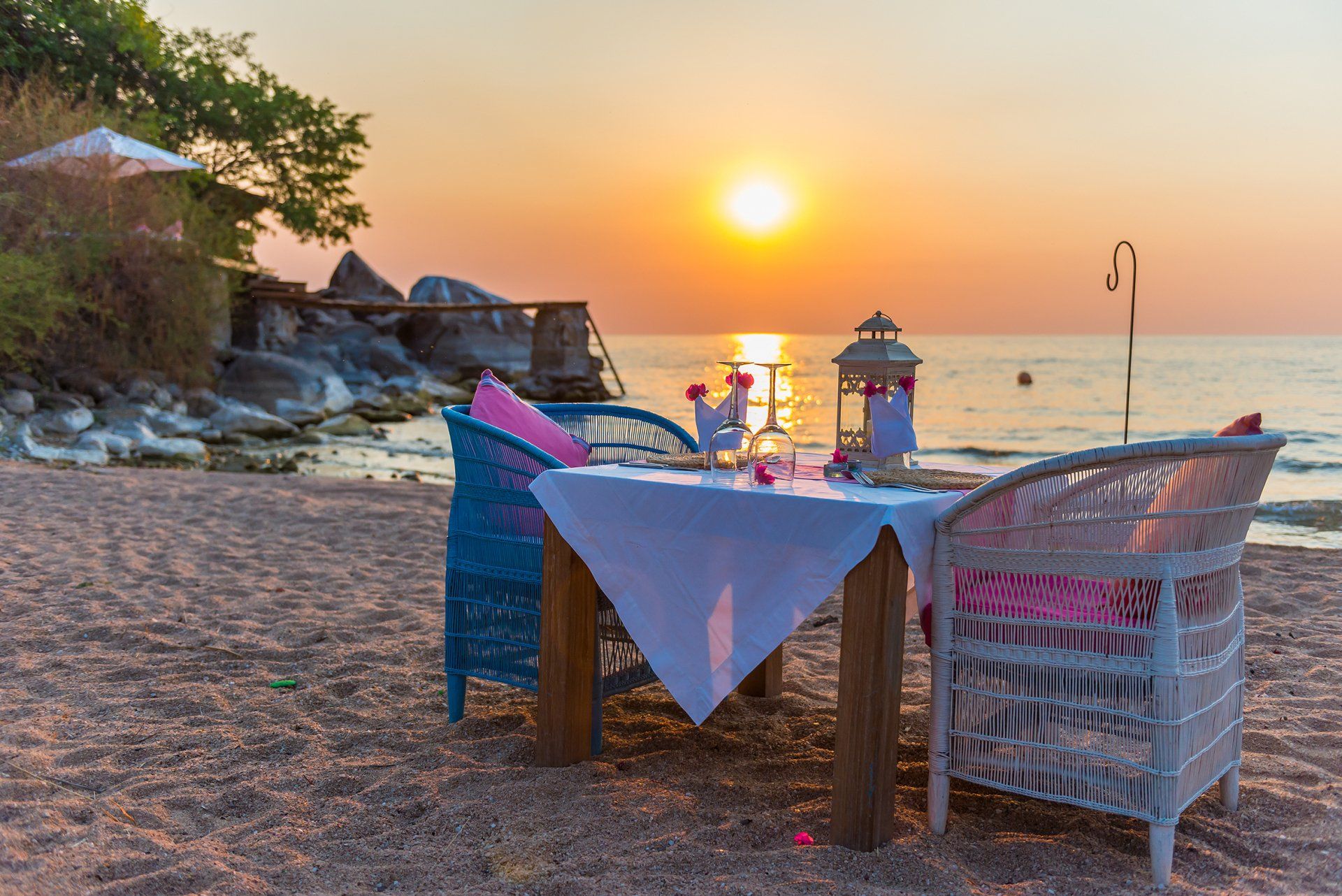 A table set for two on a sandy beach at sunset, with wicker chairs facing the ocean and rocky shoreline.