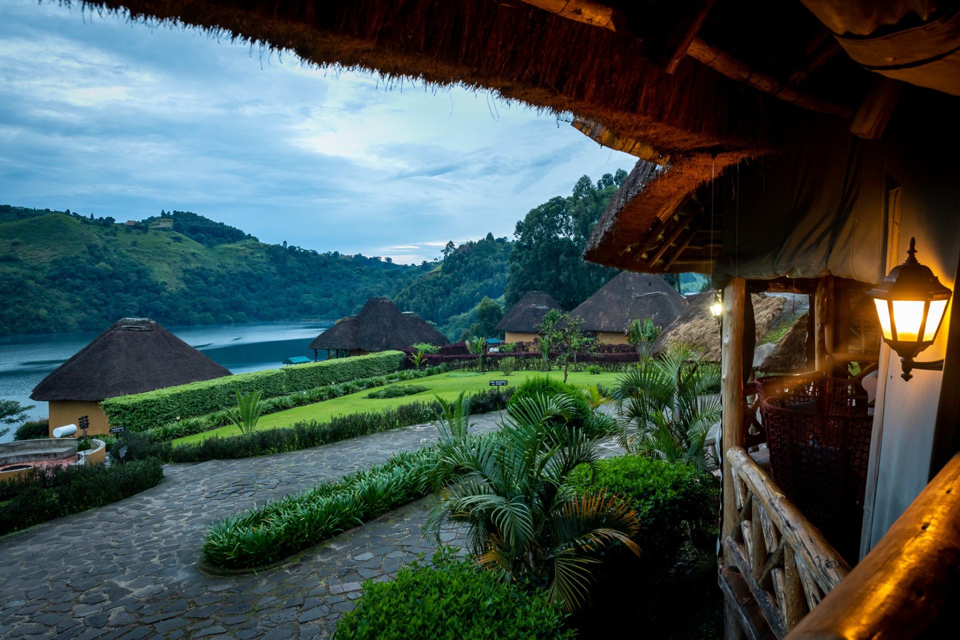 A view from a thatched-roof porch overlooking a lush garden, grass-roofed huts, and a calm lake at twilight.