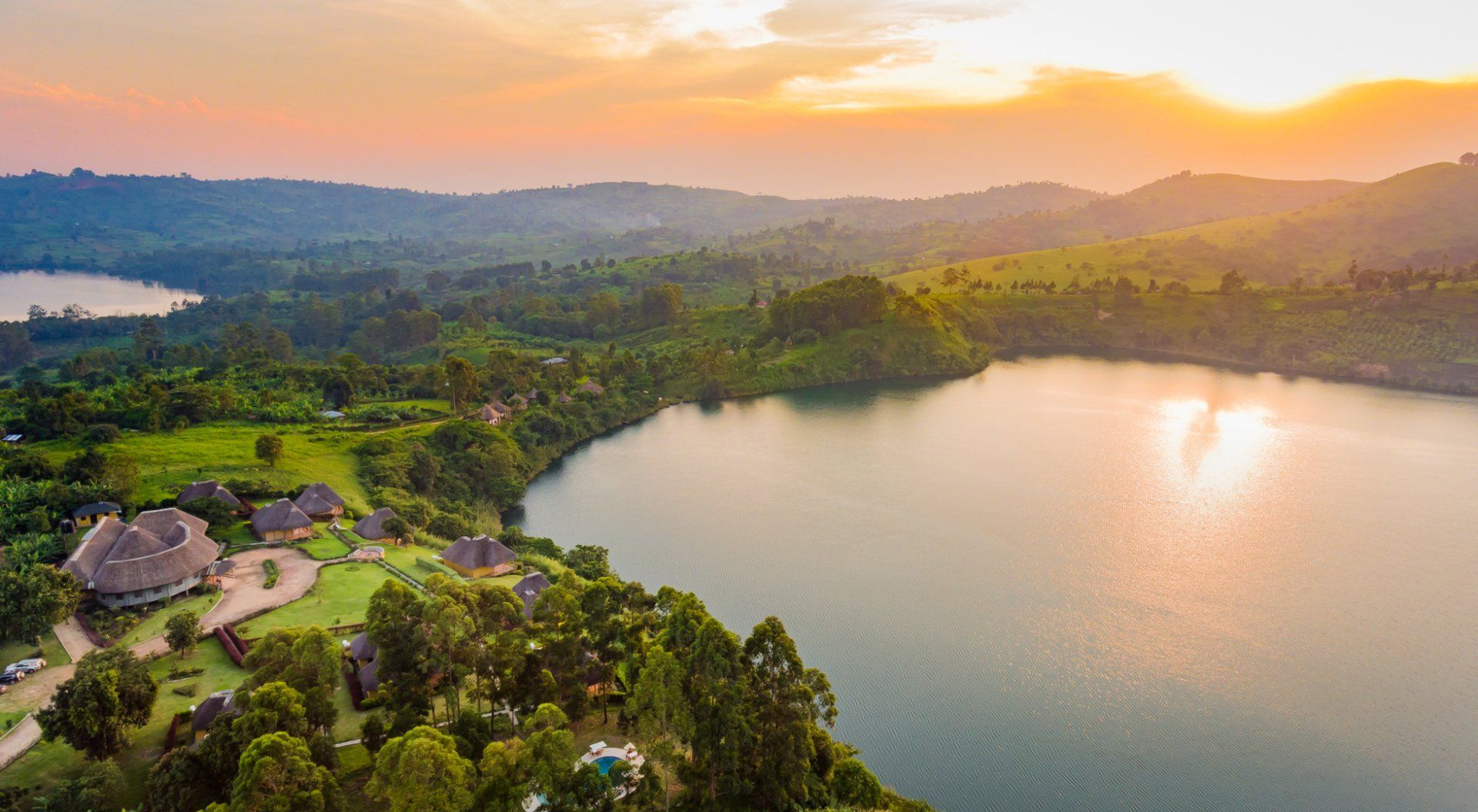 A resort with thatched-roof buildings sits on a lush green hill overlooking a calm lake at sunrise.
