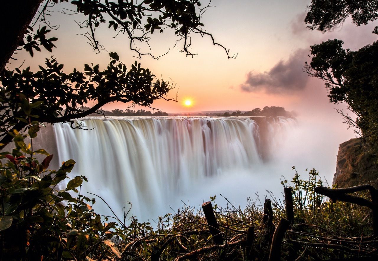 Victoria Falls framed by lush trees at sunset, with golden light reflecting over the cascading water and mist.