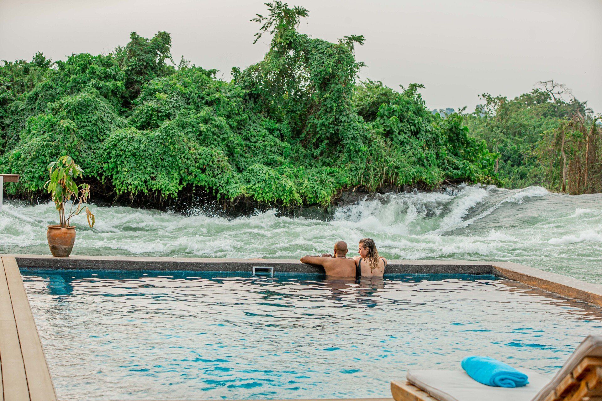 A couple relaxes in an infinity pool overlooking a river with rushing rapids and dense green forest in the background.