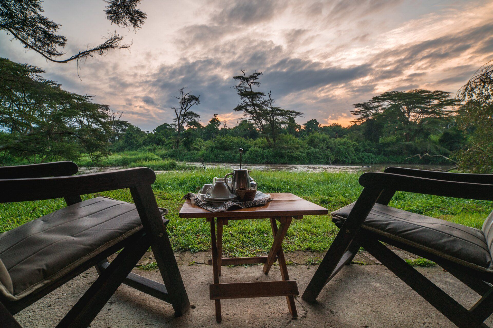 Two wooden chairs face a small table with tea service, overlooking a lush green riverbank at sunset.