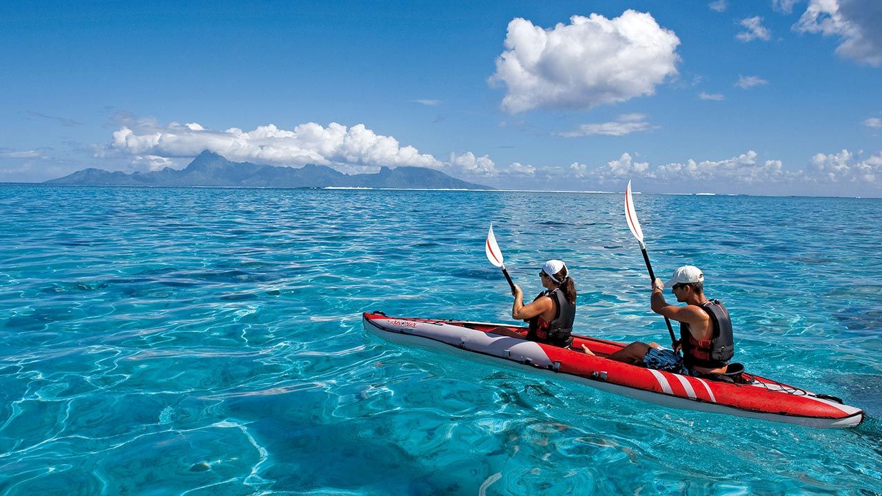 Two people paddling a red tandem kayak across clear turquoise water with a distant mountainous island in the background.