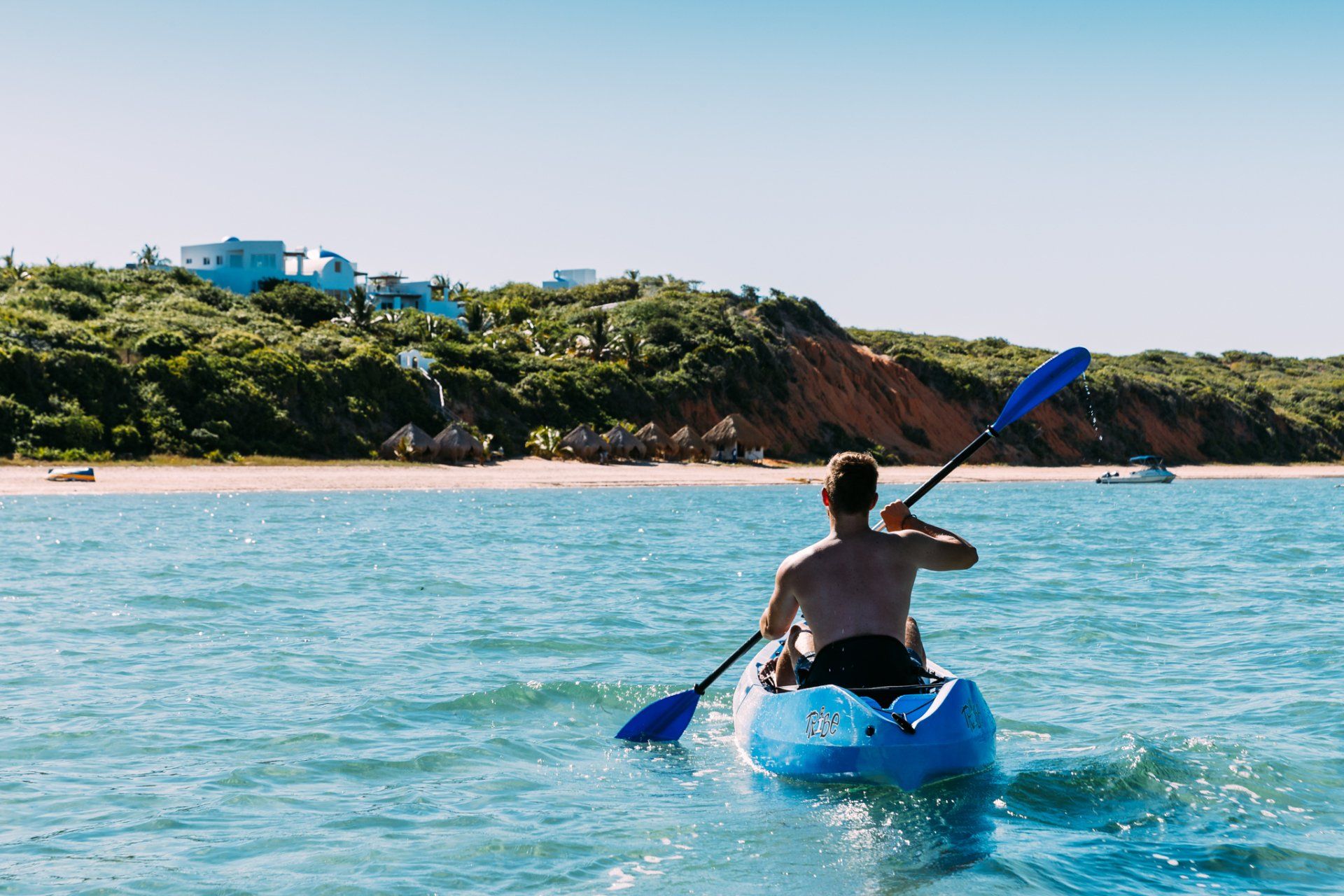 A person paddles a blue kayak across sunny turquoise water toward a sandy beach and a cliffside with vegetation.