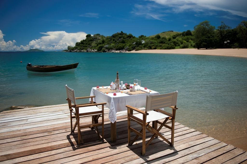 A white-clothed dining table with two folding chairs set on a wooden deck overlooking a tropical beach and blue water.
