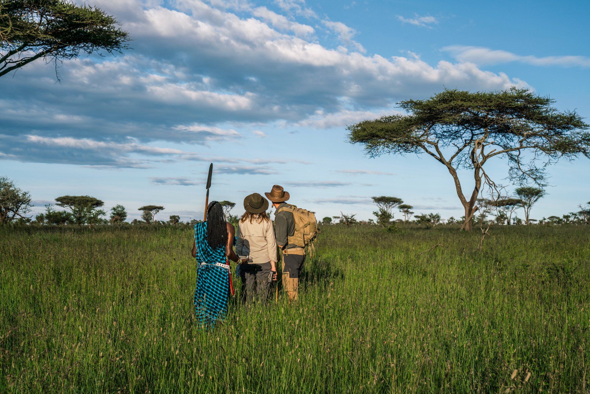 Three people walk through a lush, green savanna under a bright, partly cloudy sky, with acacia trees in the distance.