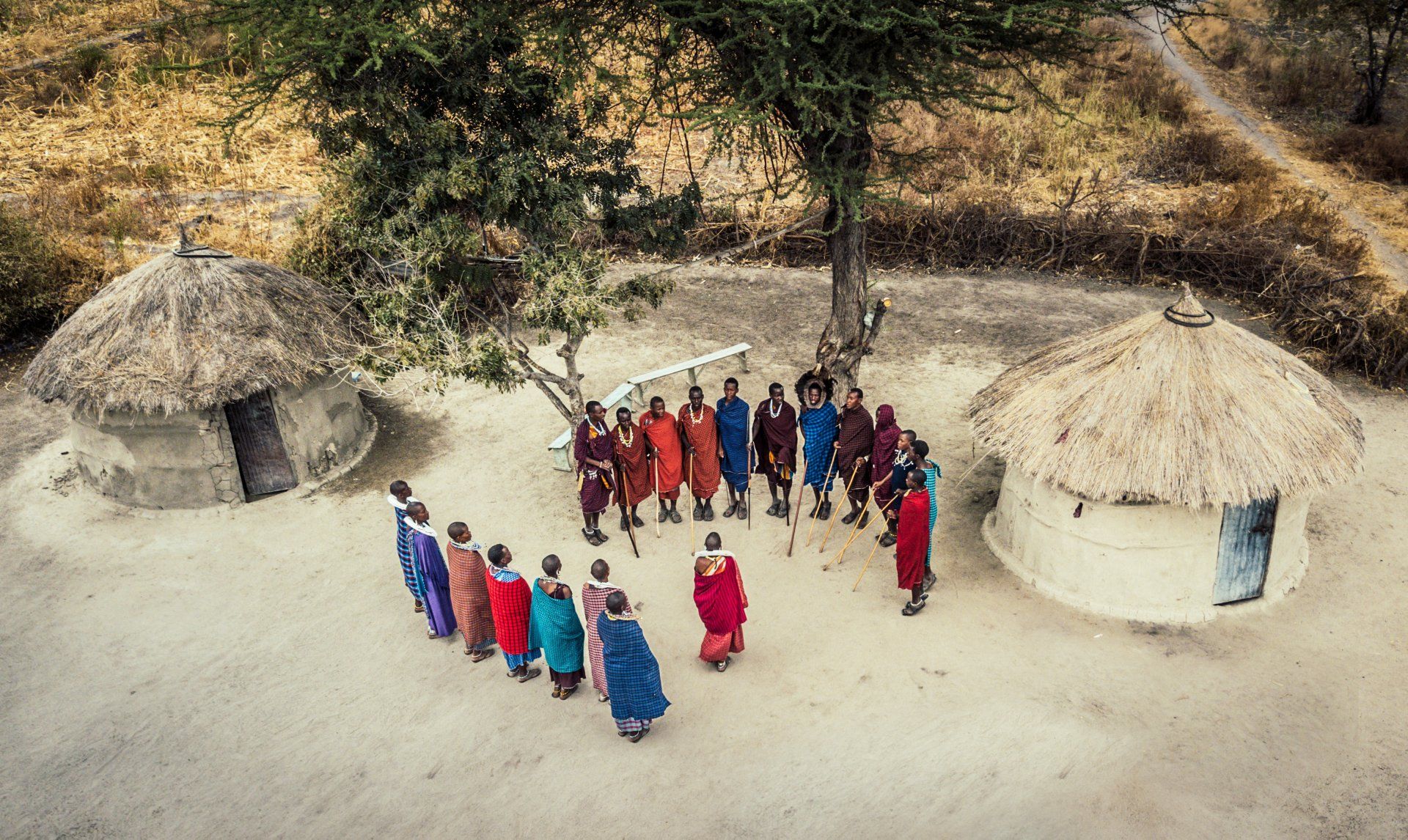 A group of people wearing colorful traditional clothing stand in a circle on sandy ground between two round thatched huts.