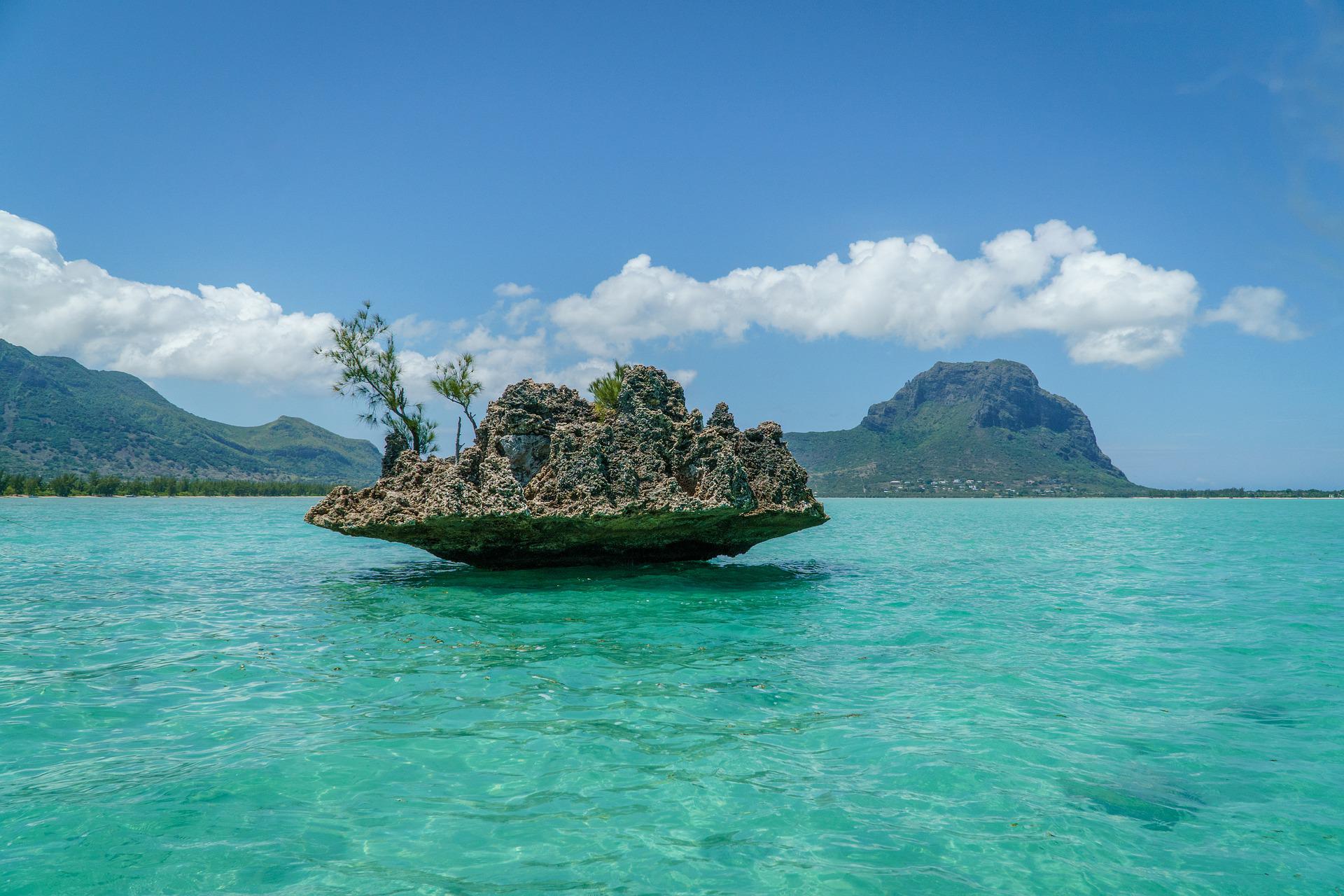 A small, rocky island with sparse vegetation floating in clear, turquoise waters, with Le Morne Brabant in the background.