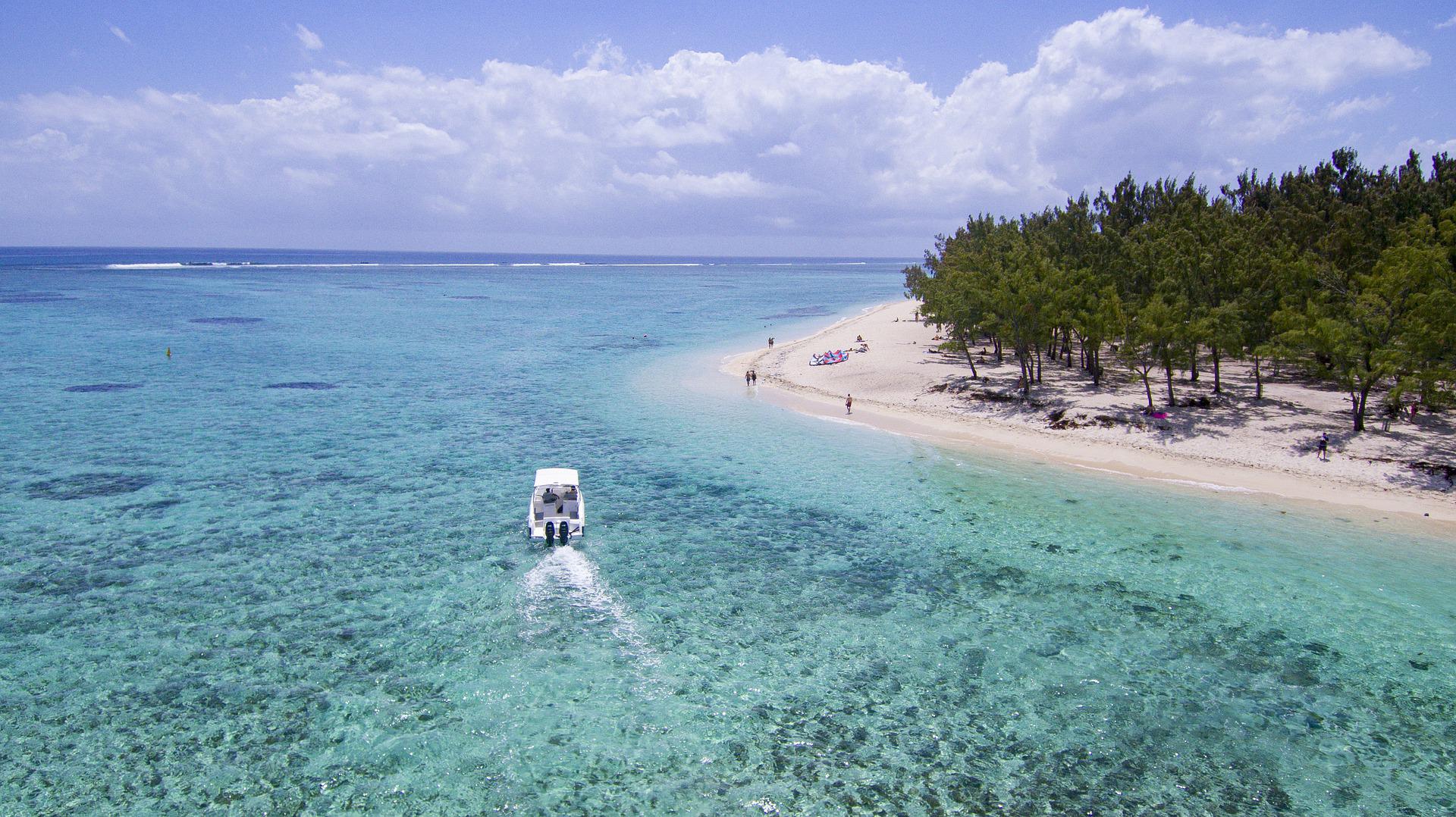 A small boat cruises through clear turquoise water alongside a sandy beach lined with green trees under a blue sky.