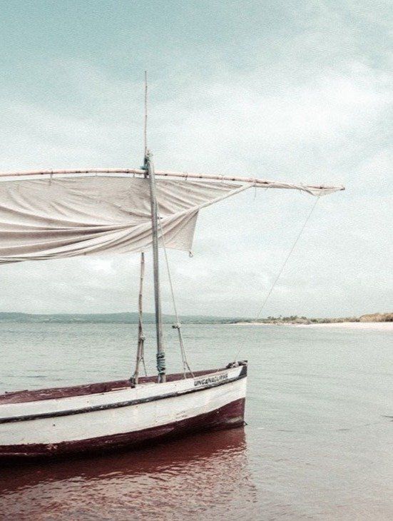 A small, white sailboat with a weathered sail rests in calm, shallow water near a light-colored sandy shore.