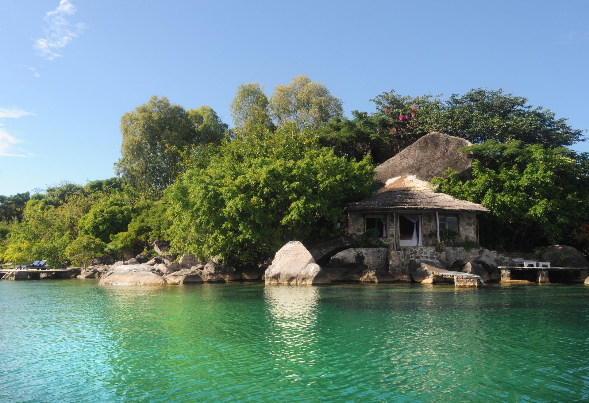 A rustic cottage nestled among lush trees and large rocks on the edge of a clear, turquoise lake under a bright blue sky.