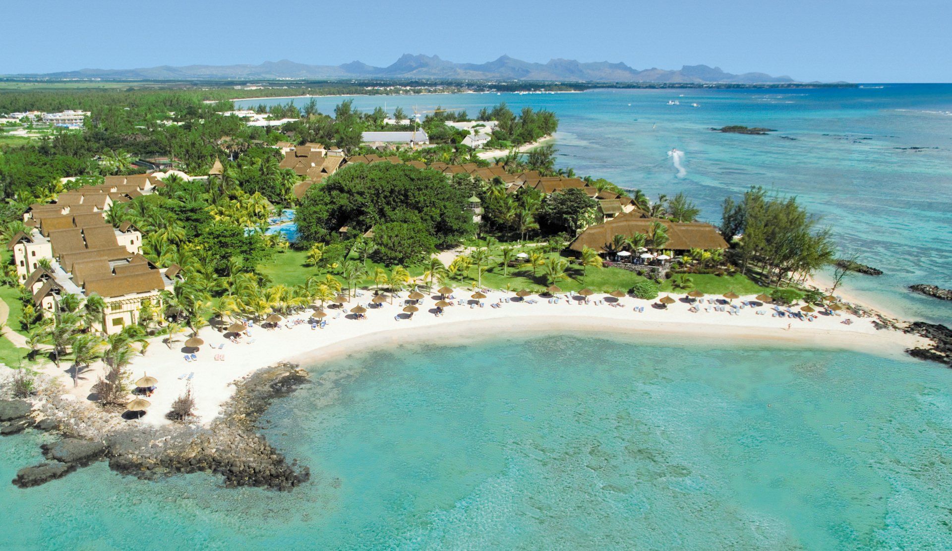 Aerial view of a coastal tropical resort with buildings, lush green trees, a white sand beach, and clear blue water.