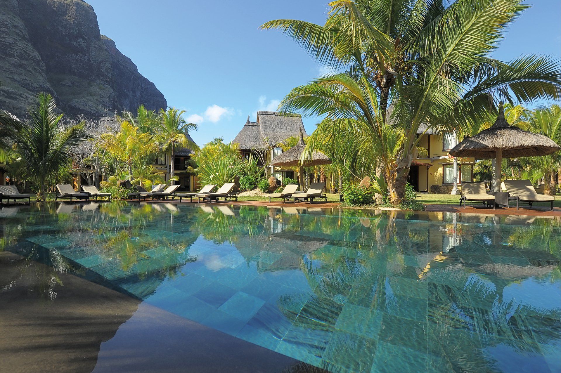 A luxurious resort pool with palm trees, thatched-roof buildings, and a large mountain in the background under a blue sky.