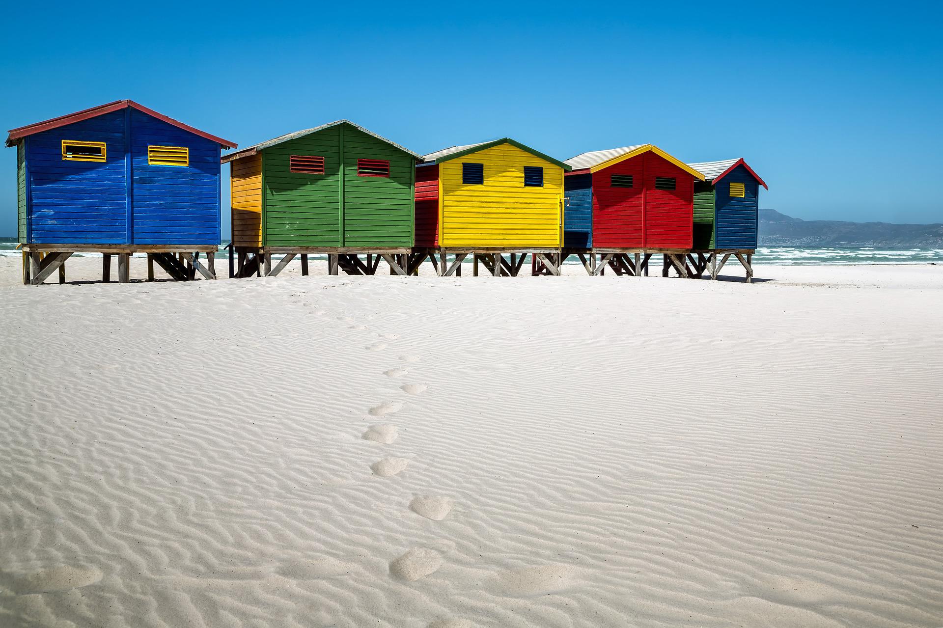 A row of colorful beach huts in blue, green, yellow, and red stands on white sand under a clear blue sky.
