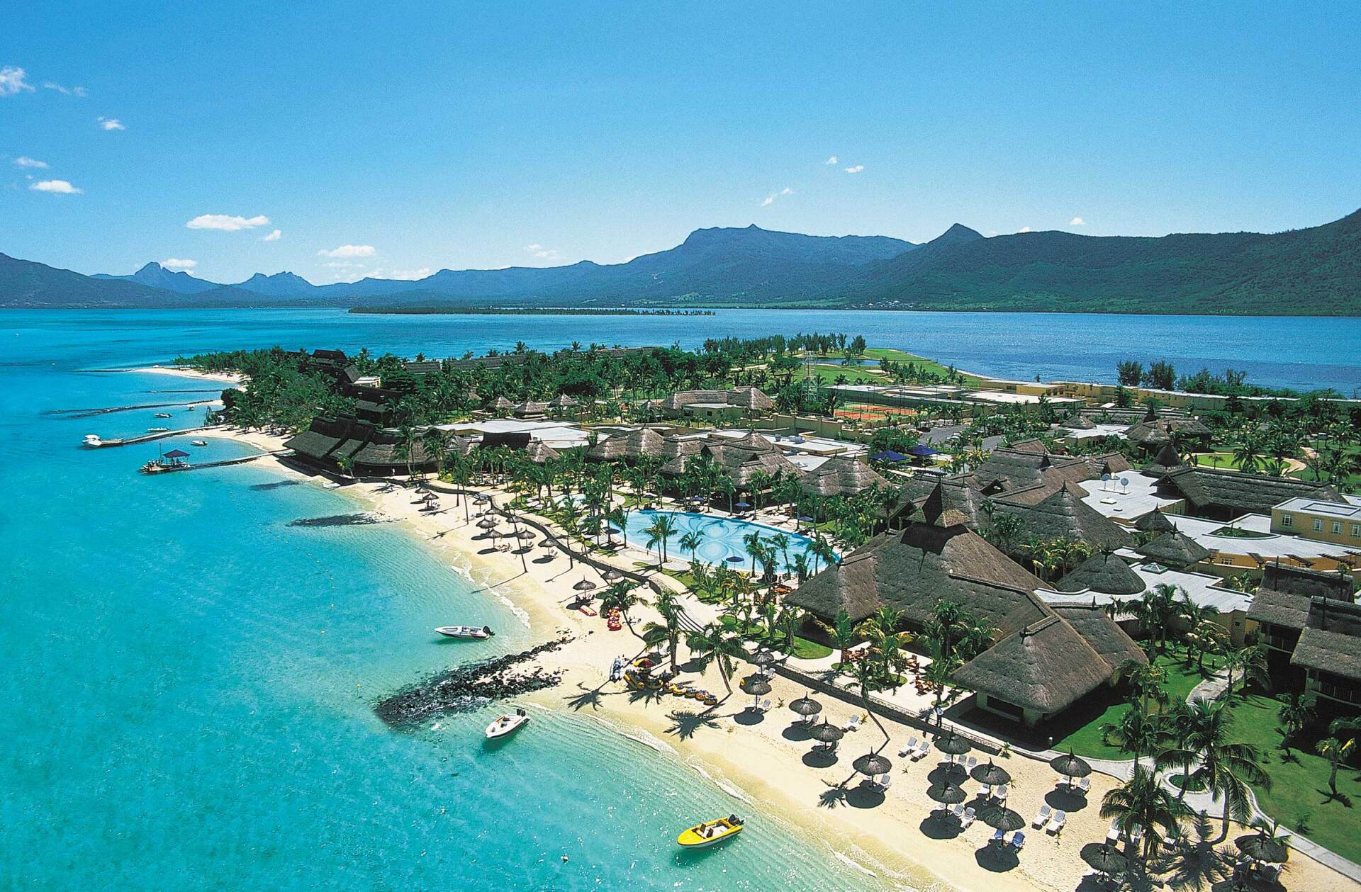 Aerial view of a tropical island resort with a sandy beach, palm trees, buildings, and turquoise water under a blue sky.