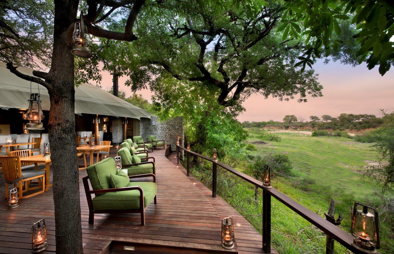 A wooden deck with green armchairs overlooks a grassy savanna at sunset, lit by lanterns under large trees.