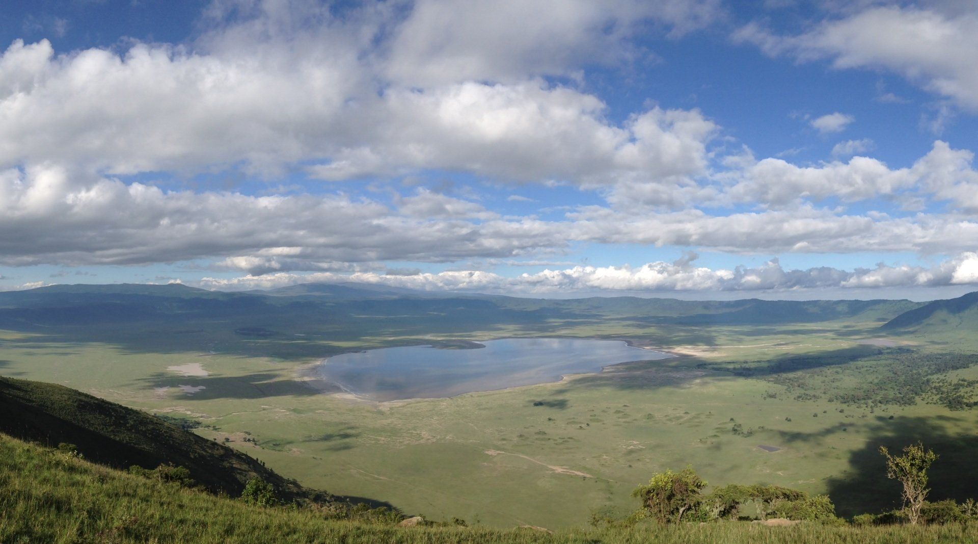 A panoramic view of a lush green crater basin with a central lake under a bright blue sky filled with white clouds.