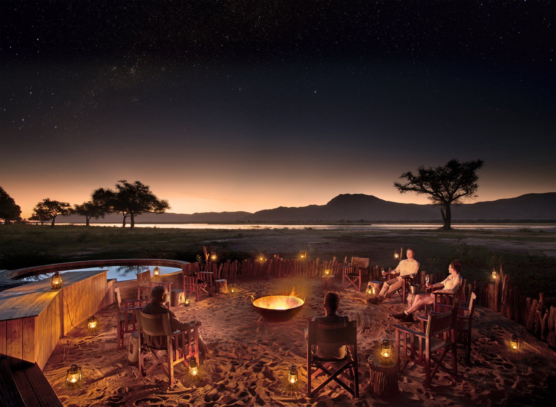 People sit around a glowing campfire in a sandy, outdoor enclosure at twilight, beneath a vast, star-filled night sky.