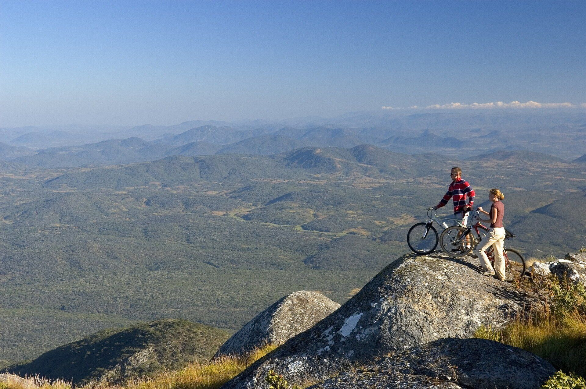 Two cyclists standing with their bikes on a rocky mountain peak overlooking a vast, rolling landscape under a clear sky.