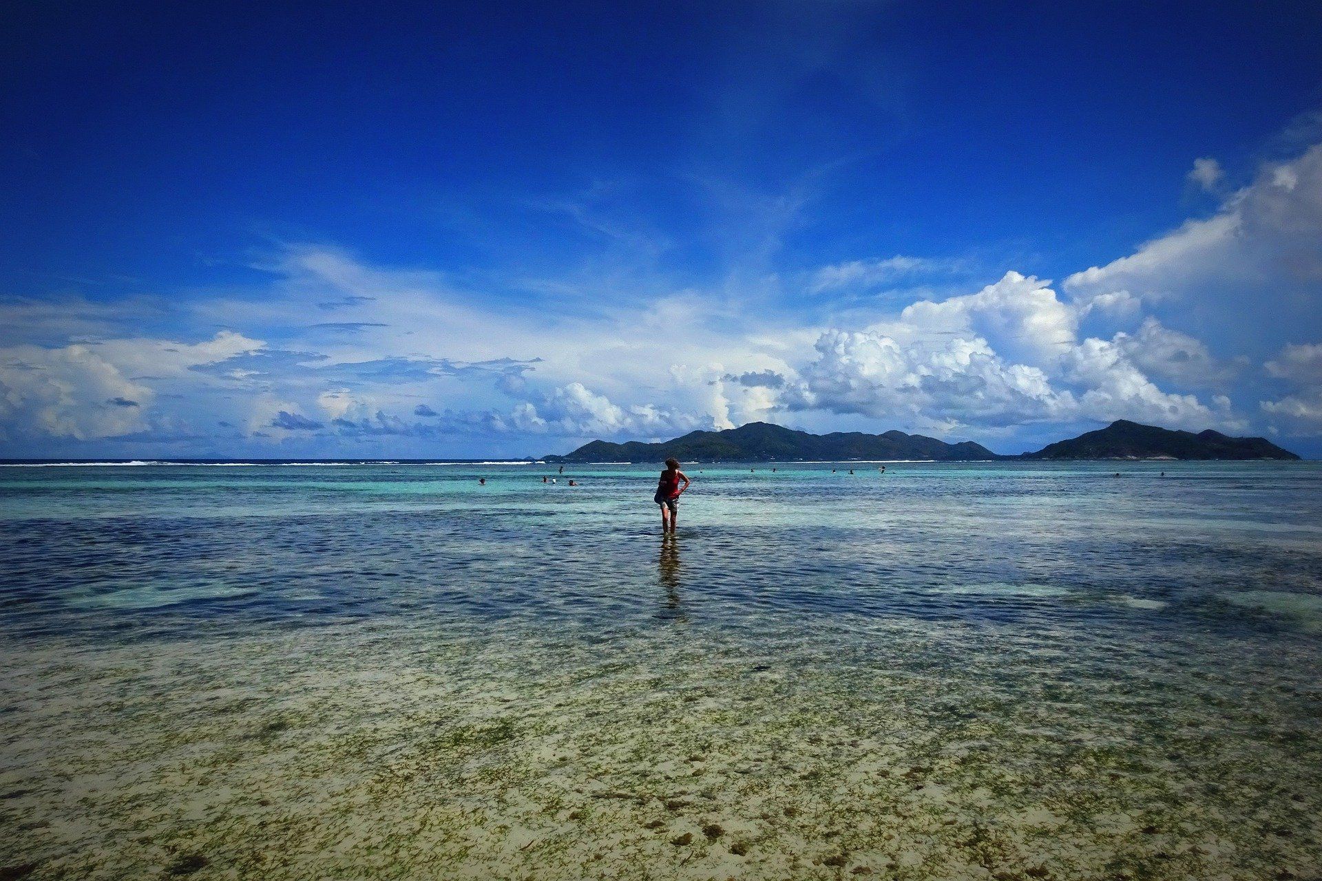 A lone person walks through shallow, clear tropical water towards distant islands under a bright, blue, cloudy sky.