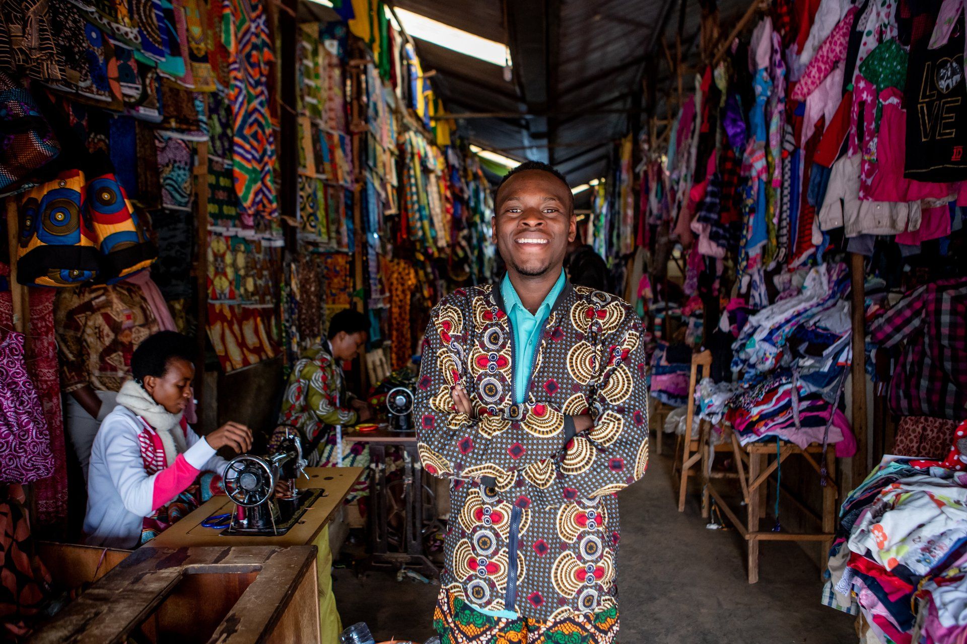 A person smiles while standing in a textile shop filled with colorful hanging fabrics and a sewing machine operator.