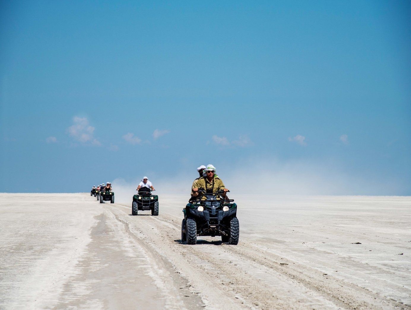 A group of people riding ATVs across a vast, dusty white desert landscape under a clear blue sky.