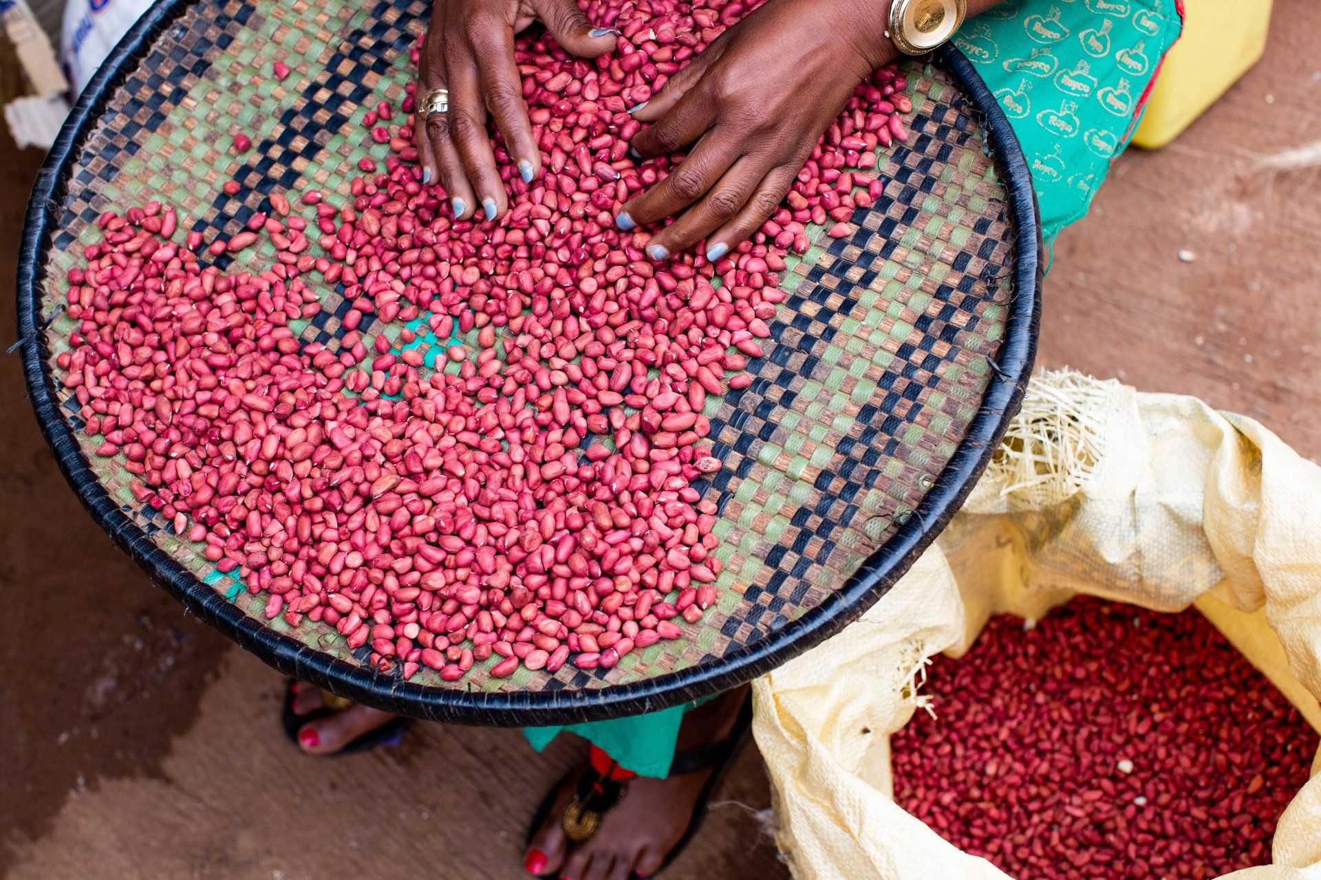A person sifts red peanuts on a woven tray, with more peanuts in a nearby bag.