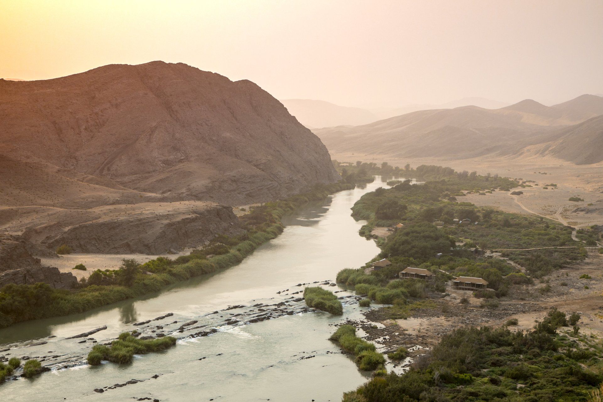 A wide river flows through a desert valley flanked by rugged mountains under a warm, hazy sky.
