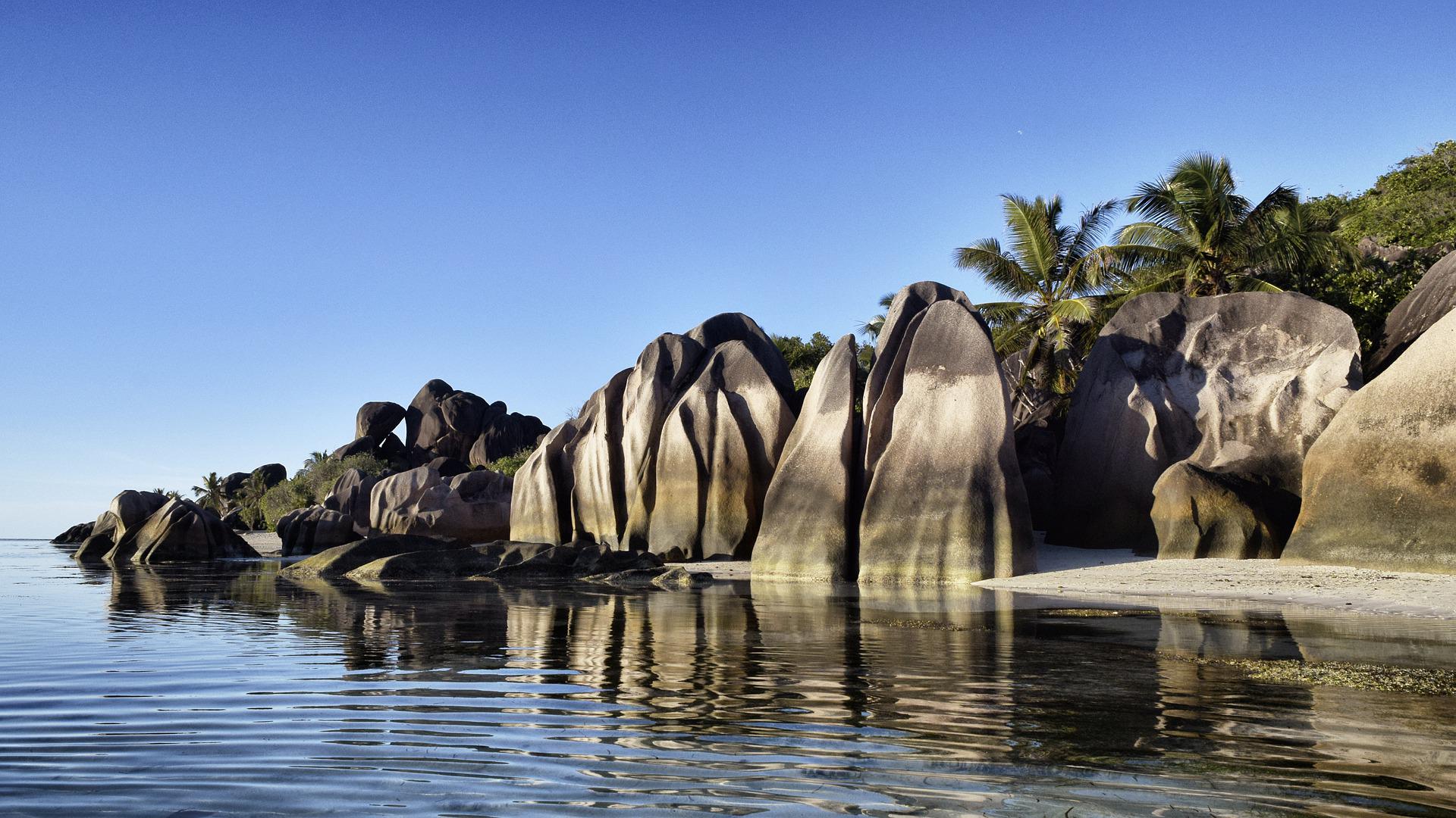 Large granite boulders rise from a calm, turquoise sea onto a sandy shore with palm trees under a clear blue sky.