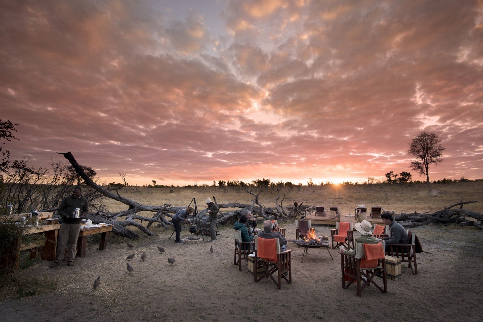A group sits in chairs around a campfire at dusk in a dry, open landscape under a cloudy, orange-toned sunset sky.
