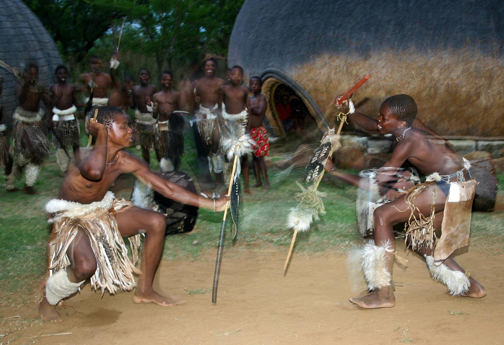 Two people perform a traditional dance with staffs in front of large, dome-shaped, thatched dwellings.
