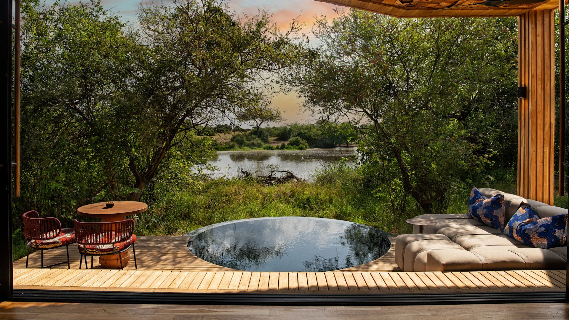 A luxury lodge terrace with a circular plunge pool, wooden deck, seating, and a view of a tranquil river and trees.