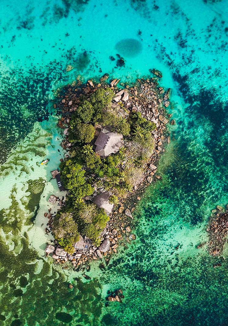 Aerial view of a small, lush green island with several huts, surrounded by vibrant turquoise and deep blue ocean water.