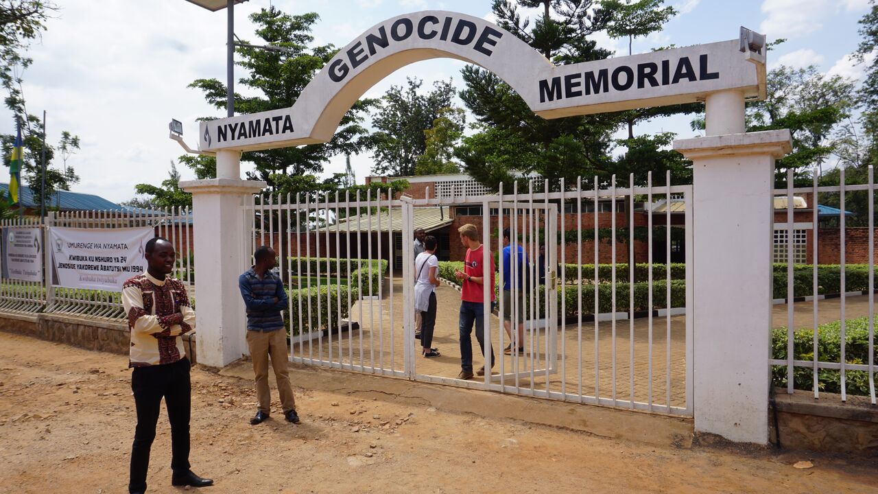 The white gated entrance to the Nyamata Genocide Memorial, with people standing near the entry in a paved courtyard.