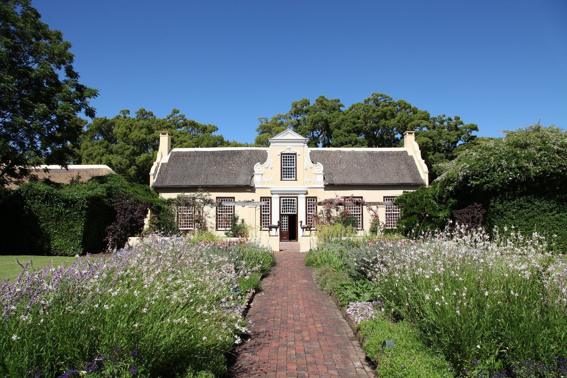 A light-colored house with a thatched roof and a central gable, framed by trees, stands behind a brick path and garden.
