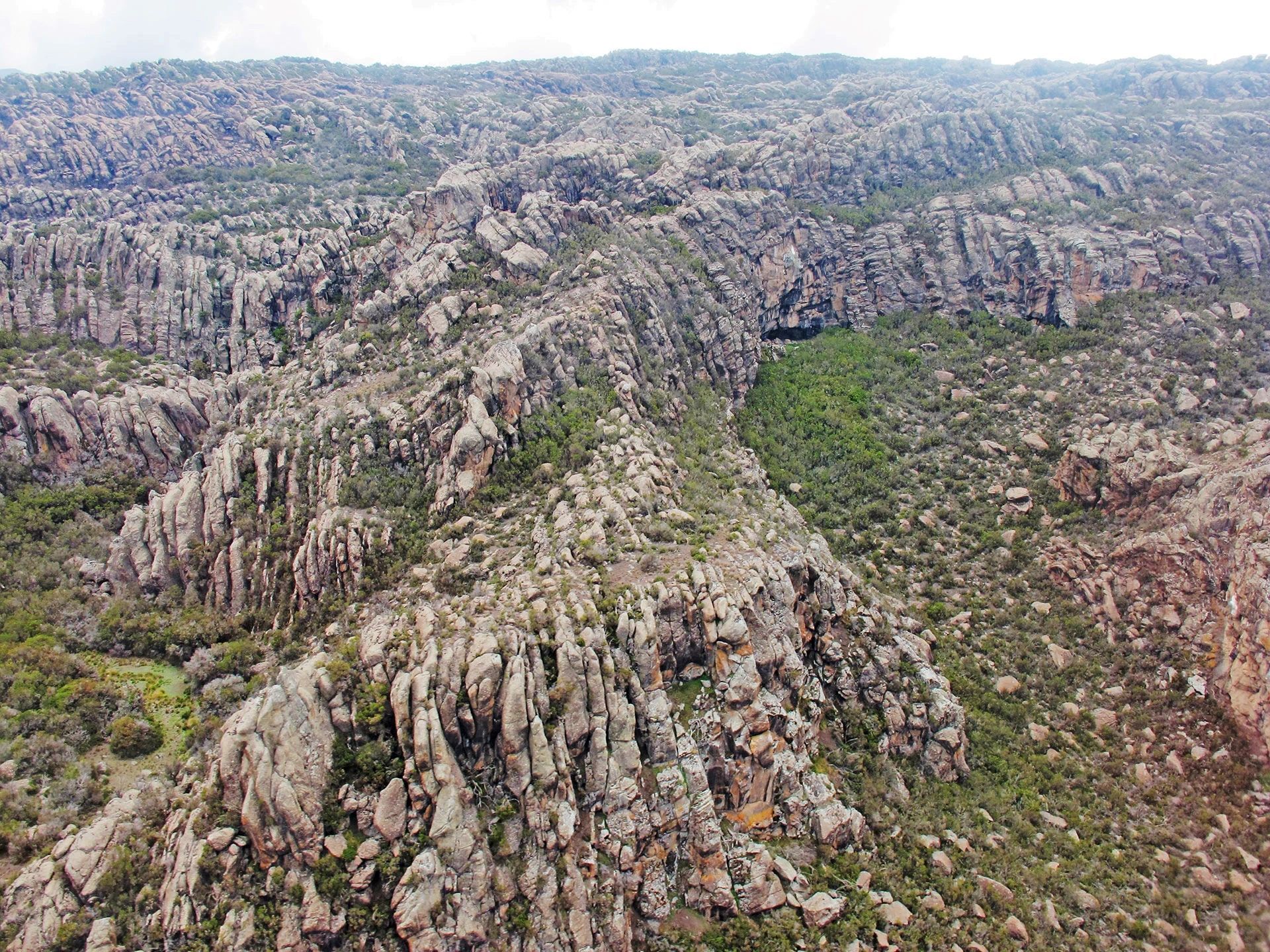 An aerial view of a rugged, rocky landscape with steep cliffs, textured stone formations, and patches of green vegetation.
