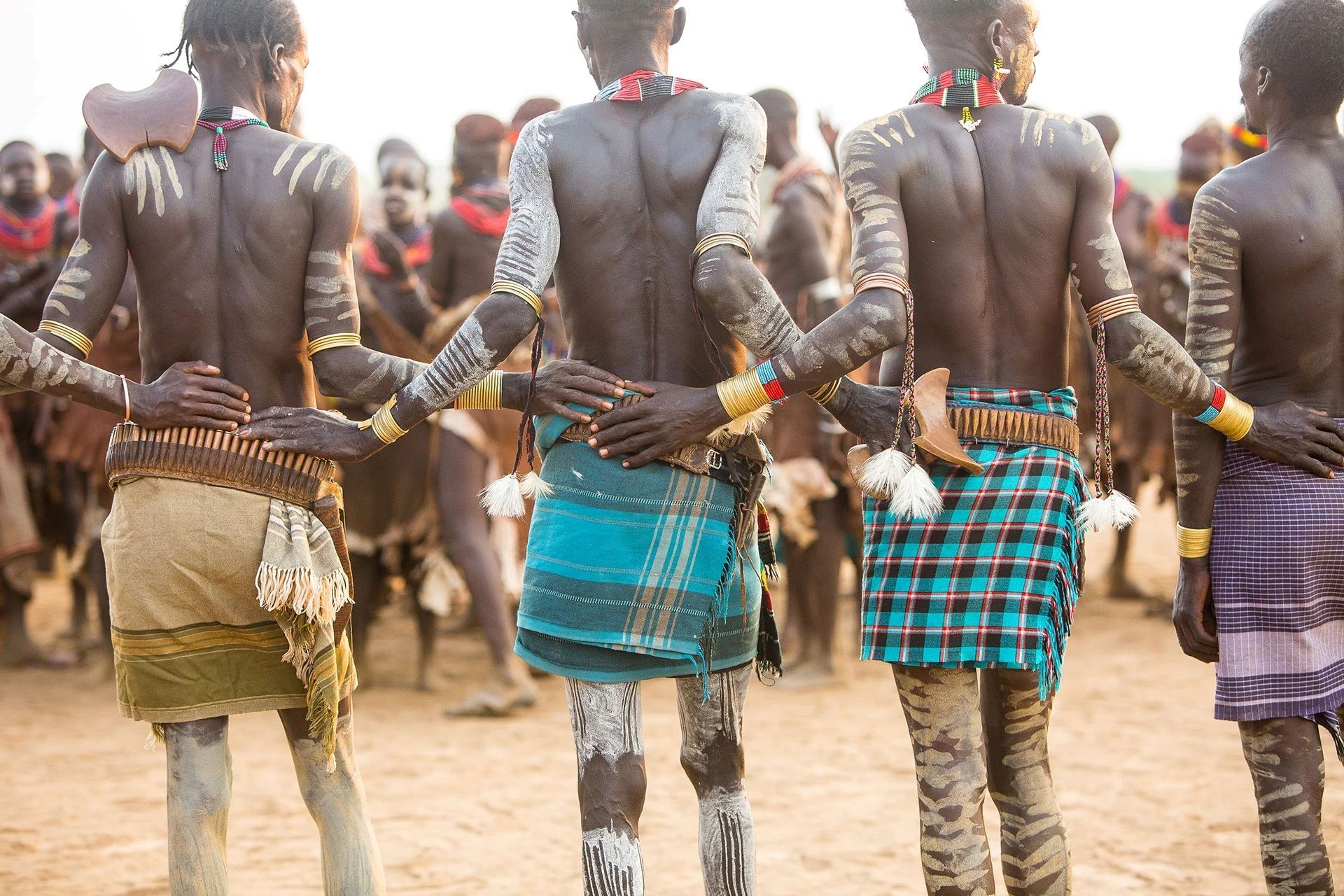 People in traditional attire with body paint standing in a line outdoors, arms linked in a dance formation.